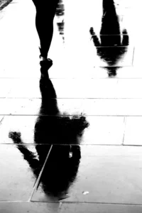 Silhouetted figures walking on a wet pavement, their dark reflections stretched and distorted in the glossy surface, creating a high-contrast, abstract scene.