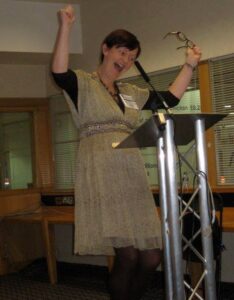 Photo of Orna at a lectern waving arms enthusiastically