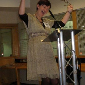 Photo Of Orna At A Lectern Waving Arms Enthusiastically