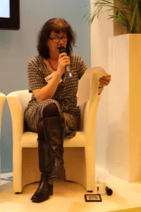 Debbie Young seated, speaking into a microphone, reading from a script