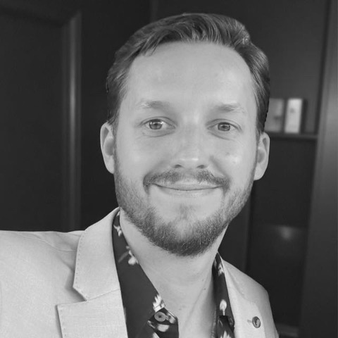 A person smiling, wearing a light-coloured blazer over a patterned shirt, with a blurred background.